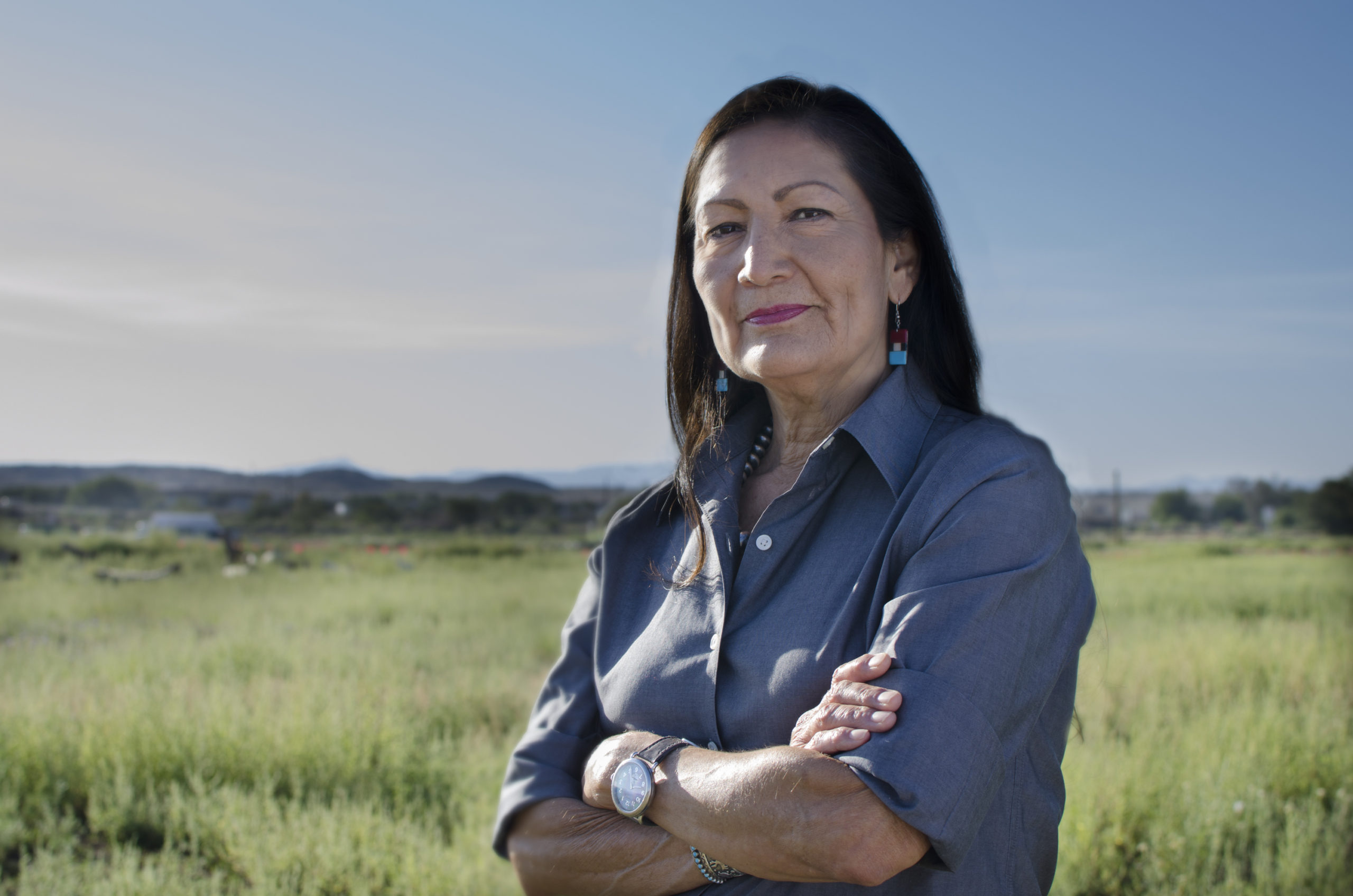 一位深色头发、戴着绿松石耳环的新墨西哥妇女双臂交叉，在开阔的田野里看着摄像机。