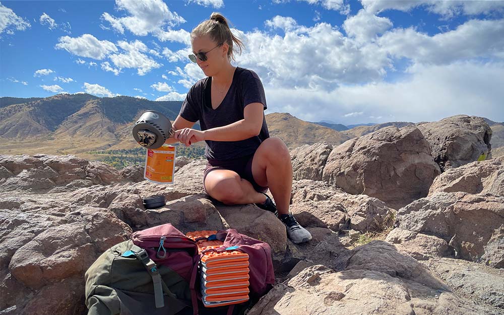 一位妇女正往一个装着脱水食物的橙色袋子里倒水，她坐在一块岩石上俯瞰着群山
